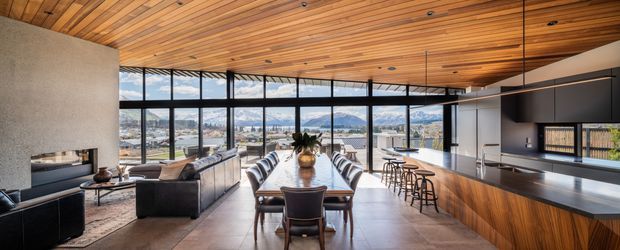 A spacious open-plan contemporary interior combining a living room, dining area, and kitchen. Full-height black-framed glazing provides expansive views toward a mountainous urban landscape. The room features a warm timber-lined ceiling, polished concrete or stone flooring, dark kitchen cabinetry, a large island with bar seating, a long timber dining table, and a living area with a fireplace and sectional seating.