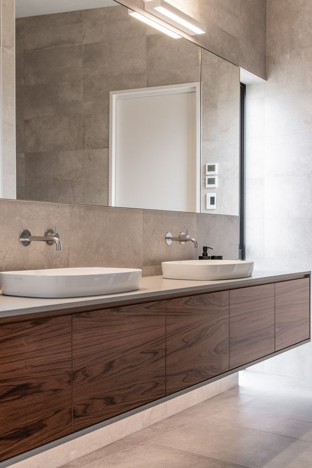 Contemporary bathroom vanity with two white vessel basins, wall-mounted chrome taps, and extensive dark timber floating cabinetry. Large-format taupe-grey stone-look wall and floor tiles, a full-width mirror, and linear wall lighting create a refined, warm modern interior.