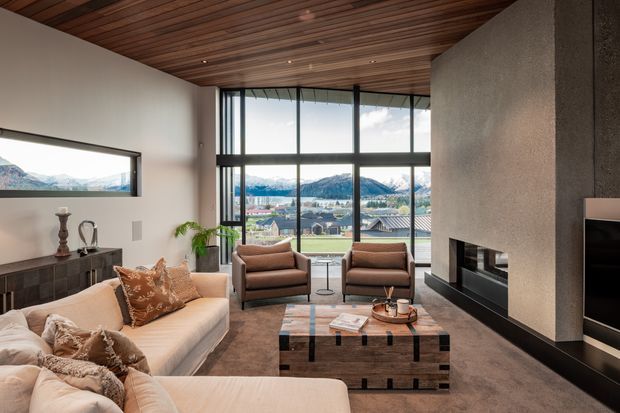 A refined, spacious living room with a high timber-lined ceiling and full-height black-framed glazing overlooking a mountainous landscape. The room features a large cream sectional sofa, two brown upholstered armchairs, a dark geometric coffee table, a textured plaster feature wall with a linear fireplace, dark built-in cabinetry, and a wall-mounted television. The overall scheme is warm, earthy, and contemporary with strong indoor-outdoor visual connection.