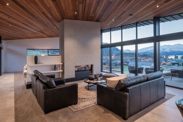 A refined contemporary living room with a central textured fireplace volume, leather sofas and armchairs arranged around a low coffee table, and an open media shelving unit. Warm timber lining covers the ceiling, while expansive black-framed glazing opens to a balcony or terrace and provides views toward a mountainous townscape. Recessed ceiling lighting, neutral finishes, and a large area rug create a comfortable, sophisticated interior.