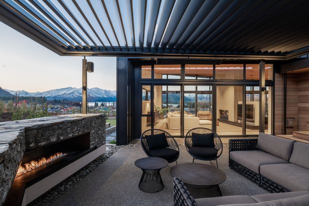 A sophisticated covered outdoor lounge terrace with a linear open-flame fireplace, dark stone masonry, contemporary black-framed glazing, and a louvred overhead roof. The space is furnished with black lounge chairs, woven sofas, and round coffee tables, and opens toward a broad mountain and suburban view.