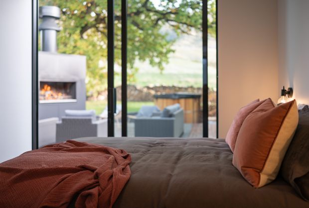 A warm, contemporary bedroom with a neatly made bed in muted brown bedding and layered rust, tan, and dark cushions. Full-height black-framed glazing opens the room visually to an outdoor seating area and fireplace, while a small wall-mounted bedside light adds a refined, cozy accent.