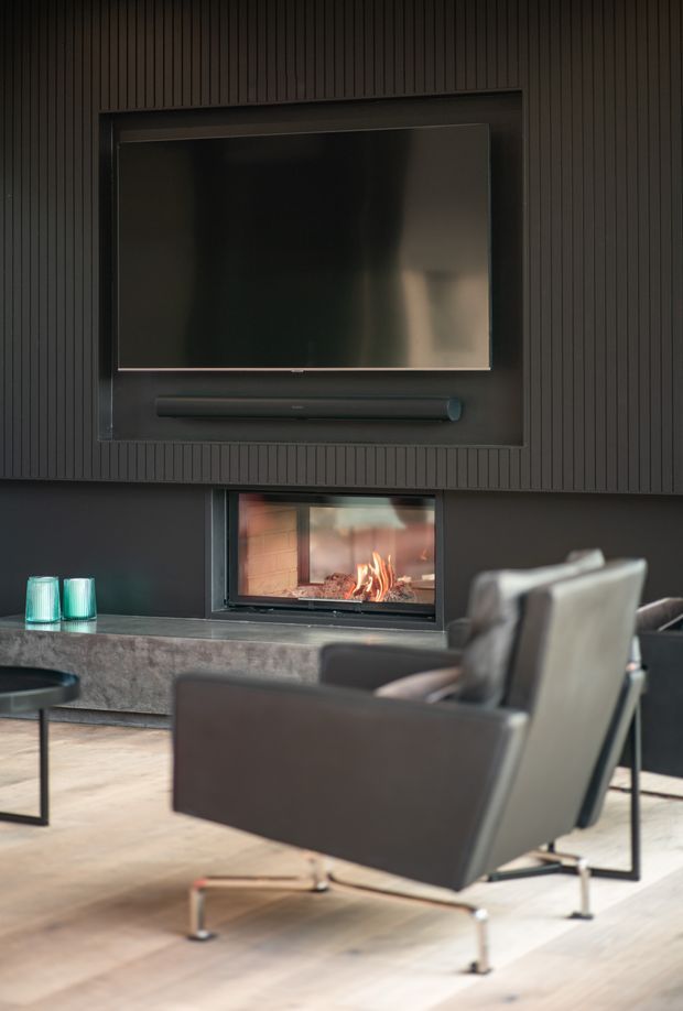 A moody contemporary living room centred on a dark media wall with a wall-mounted television and soundbar above a built-in fireplace. Vertical dark wall panelling, a stone hearth or low console, pale timber flooring, and a sculptural upholstered lounge chair create a refined, luxurious atmosphere.