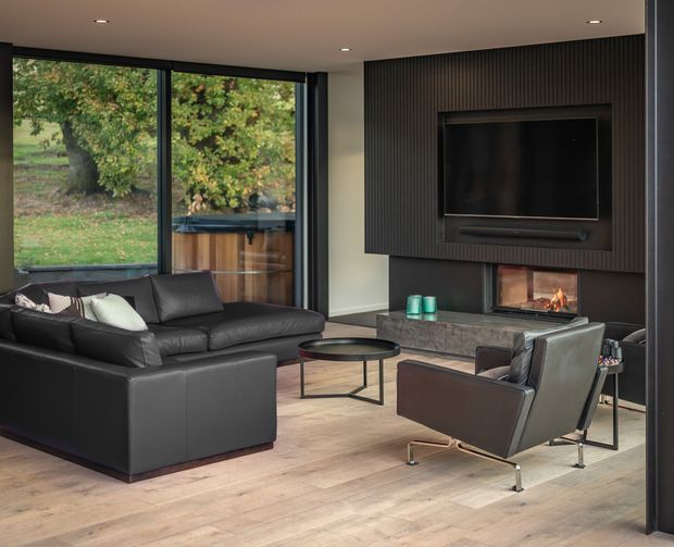 Contemporary living room with a black leather sectional sofa, modern lounge chair, round coffee table, and a wall-mounted television above a built-in fireplace. Full-height dark-framed glazing opens views to greenery outside, while a dark vertical-panel feature wall and pale timber flooring create a refined, moody interior.