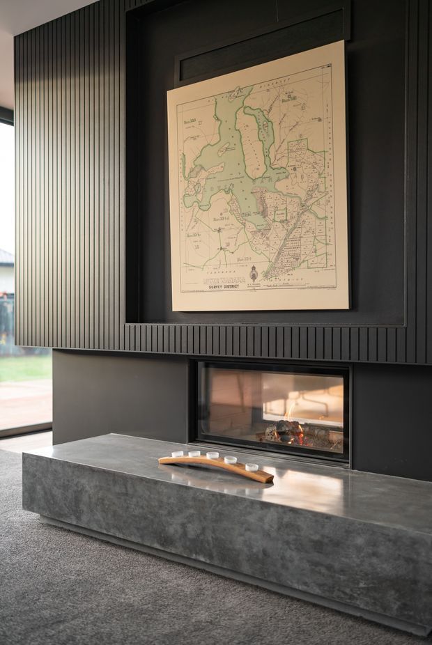 A refined contemporary living room feature wall with a built-in linear fireplace set into dark charcoal cabinetry and vertical fluted wall panelling. A framed vintage map hangs above the fireplace, while a substantial grey concrete or stone hearth projects into the room. The space has dark, moody tones, a carpeted floor, and natural light entering from glazing at the left.