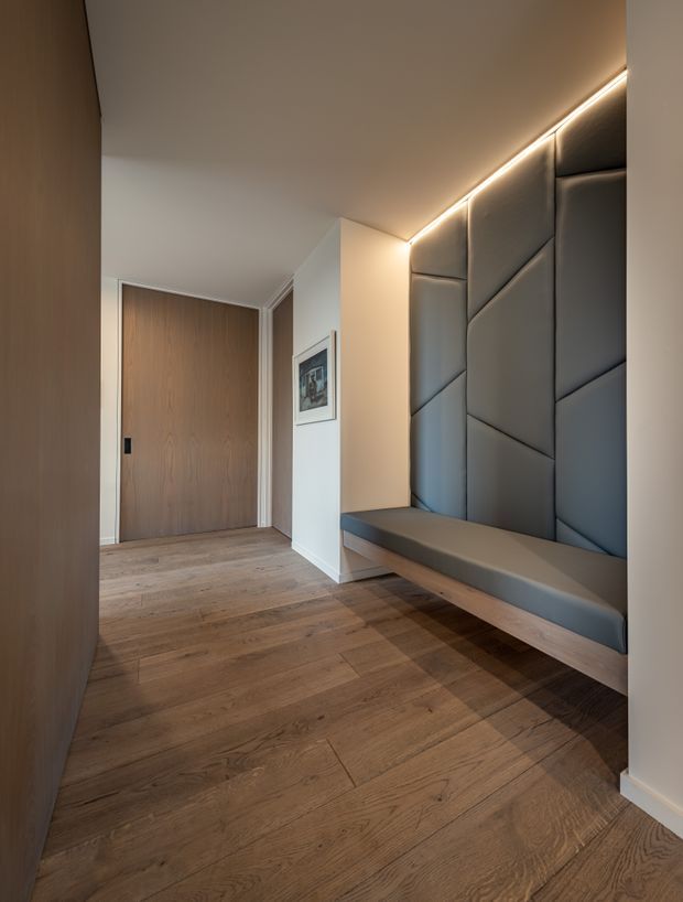 A refined modern entry hall with warm timber flooring and a timber door, featuring a built-in floating bench beneath a large geometric padded wall panel. Concealed linear lighting highlights the ceiling edge and creates a warm, softly illuminated atmosphere. White walls and restrained detailing reinforce the minimalist design.
