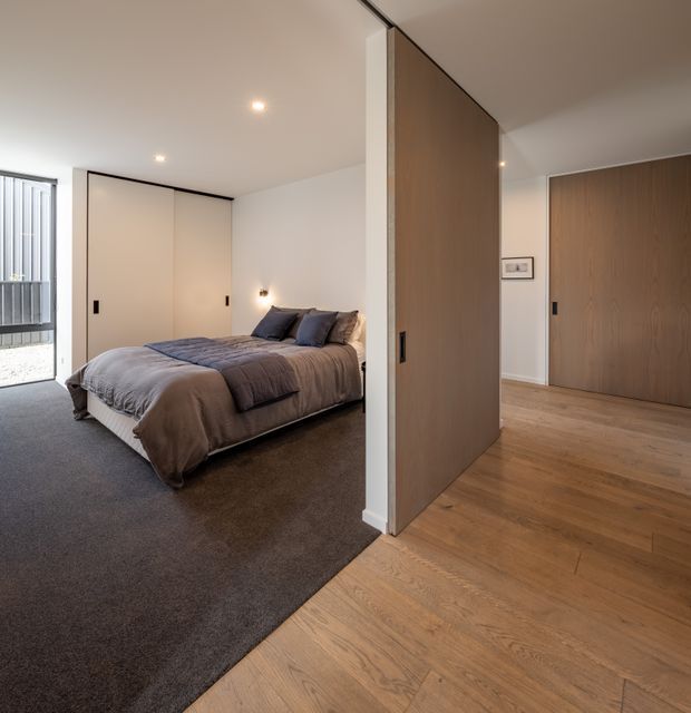 A contemporary bedroom viewed from an adjoining timber-floored hallway or entry area. The bedroom features a low upholstered bed with layered grey bedding, dark carpet, pale built-in wardrobes, recessed ceiling lights, and a small wall light. A large timber pivot or sliding partition door separates the bedroom from the hallway, which has warm oak flooring and a matching timber door. Glazing or a balcony opening is visible at the left.