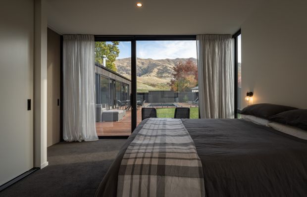 A contemporary bedroom with a large bed dressed in dark bedding and a grey plaid runner, dark carpet, recessed ceiling lighting, and full-height curtains framing expansive black-framed glazing. The doors open to a timber deck and landscaped outdoor area, with rugged mountains visible beyond.