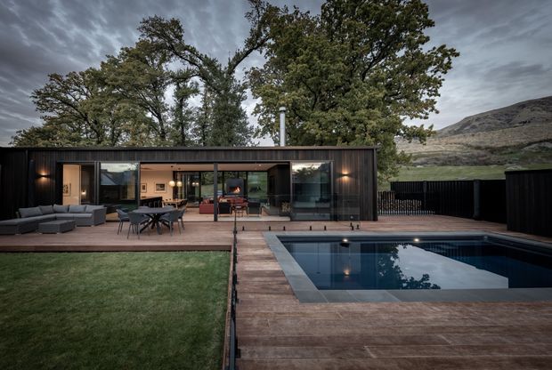 A low, single-storey contemporary residence with a dark timber-clad rectangular facade, extensive glazed sliding doors, and a warm open-plan interior. A broad timber deck connects the house to an outdoor dining and lounge area and a rectangular swimming pool. Mature trees and a rugged mountain landscape form the surrounding setting, while the overcast sky and dark finishes create a dramatic atmosphere.