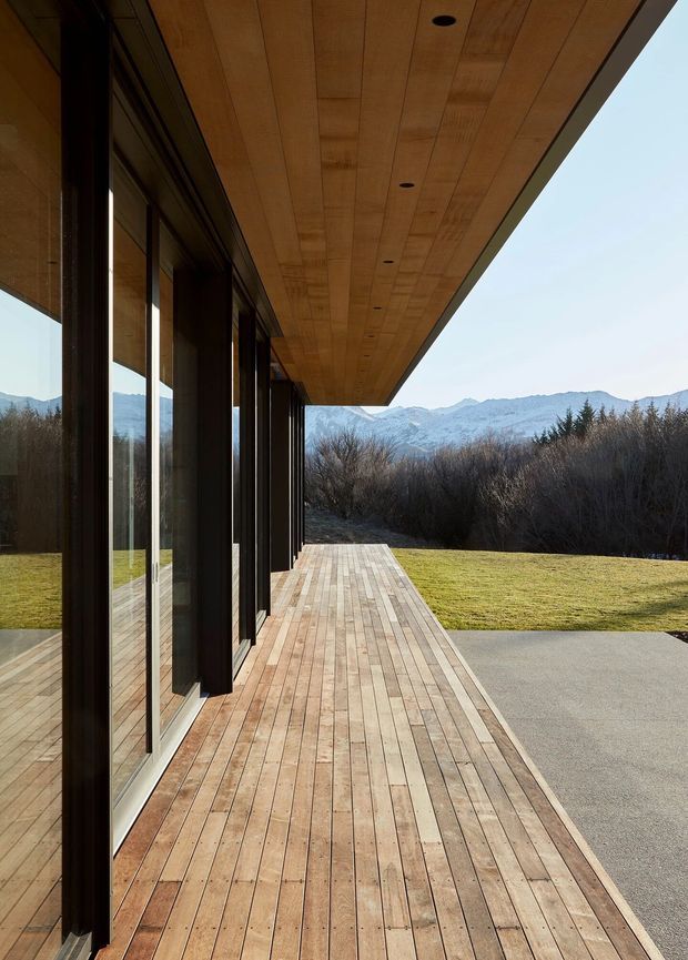A modern covered outdoor deck runs alongside a glazed building facade, with dark vertical window and door frames, extensive floor-to-ceiling glazing, and a warm timber soffit with recessed downlights. The timber decking extends toward a lawn and gravel or concrete area, with dense vegetation and snow-covered mountains visible beyond. The image emphasizes the architectural overhang, linear perspective, and strong indoor-outdoor connection.