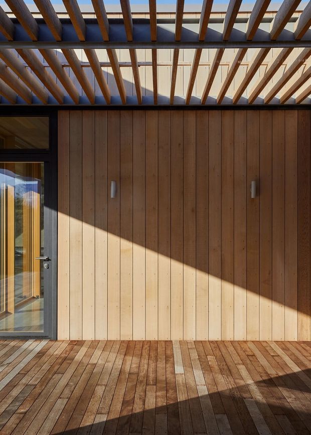 A sunlit architectural deck or terrace framed by warm timber construction. The space features vertical timber wall cladding, matching timber decking, a dark-framed glazed door at left, and an overhead pergola with exposed timber rafters casting strong linear shadows. The composition is minimal and highly geometric, with warm natural wood tones contrasted by dark metal framing.