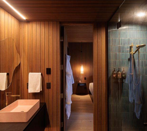 Warm, atmospheric bathroom with extensive vertical timber wall and ceiling panelling, a sculptural mirror, pale vessel basin on dark joinery, and a glass-enclosed shower lined with blue-green vertically ribbed tiles. An open doorway leads directly into a bedroom area visible in the background, with a bed and hanging robe. Integrated linear lighting and a small wall light create a refined, spa-like setting.