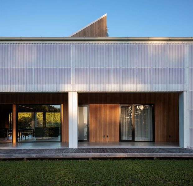 Front exterior view of a contemporary single-storey residence with a broad translucent corrugated facade screen, warm vertical timber cladding, extensive glazed openings, and a timber deck opening onto a lawn. A small pitched timber-clad roof form rises behind the main roofline.