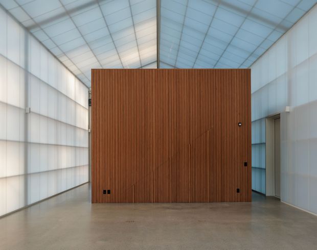 A large, minimalist interior with translucent white wall panels and a translucent pitched roof creating a bright, diffuse atmosphere. The polished concrete floor surrounds a freestanding rectangular volume clad in vertical warm timber battens, with a concealed or integrated doorway and small black hardware. The space reads as an architectural hall, gallery, or commercial interior rather than a conventional residential room.