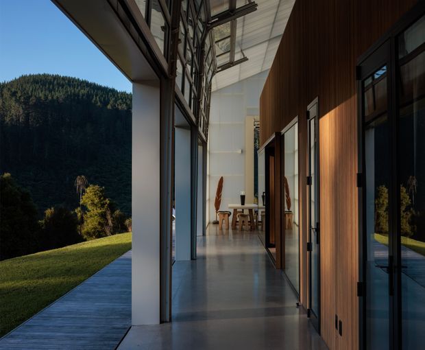 A modern interior passage or gallery beside expansive glazing, opening toward a dining area. The space features a polished concrete floor, tall dark-framed glass doors and windows, a warm vertical timber-paneled wall with integrated openings, exposed structural framing, and a high translucent ceiling. A landscaped lawn and densely forested hillside are visible outside.