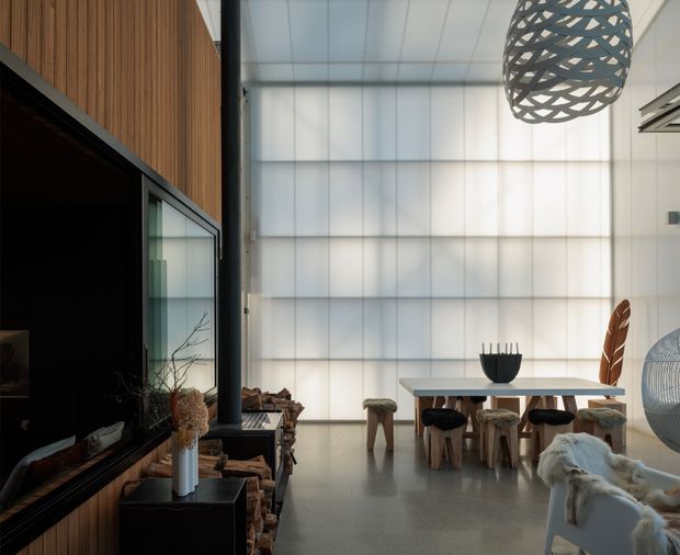 A contemporary open-plan living and dining space with a dramatic double-height translucent-panel wall, polished concrete floor, and warm vertical timber wall cladding. A large black-framed fireplace or media wall anchors the left side, while a simple dining table with rustic timber stools occupies the brighter rear area. Sculptural pendant lighting, a black structural column, and layered natural furnishings create a refined yet organic atmosphere.