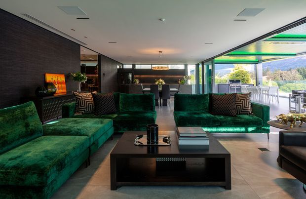 A sophisticated open-plan living room with deep emerald velvet sofas arranged around a large black coffee table. Dark wood wall panelling, a bar or kitchen area in the background, sculptural lighting, patterned cushions, and extensive floor-to-ceiling glazing create a dramatic yet refined interior. The glazing opens toward an outdoor terrace and leafy landscape beyond.