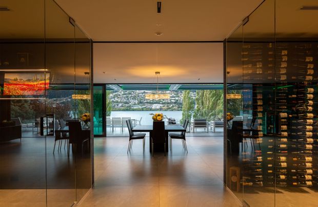 A dramatic interior view through a glass-lined passage into a contemporary dining area with a dark dining table and chairs, opening onto a waterfront terrace. A large built-in wine display and cellar occupy the right side, while mirrored or reflective glazing extends along both sides. The space features polished pale flooring, warm ambient lighting, expansive glazing, and views across the water to a populated hillside.