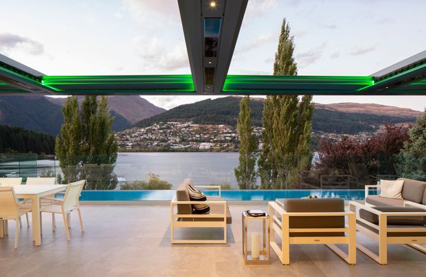 A covered outdoor terrace overlooking a lake and mountainous settlement, with a narrow infinity-edge pool along the view. The space features pale tiled flooring, light timber outdoor lounge furniture, a dining table and chairs, and a dramatic overhead canopy with integrated linear green lighting. Floor-to-ceiling glazing or open sides provide uninterrupted views of the landscape.