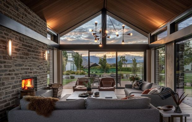 A spacious contemporary living room with a dramatic pitched timber ceiling, exposed masonry feature wall and built-in fireplace. Floor-to-ceiling glazing frames a landscaped garden and distant mountain view. A large sectional sofa, lounge chairs and sculptural pendant create a warm, refined seating area.