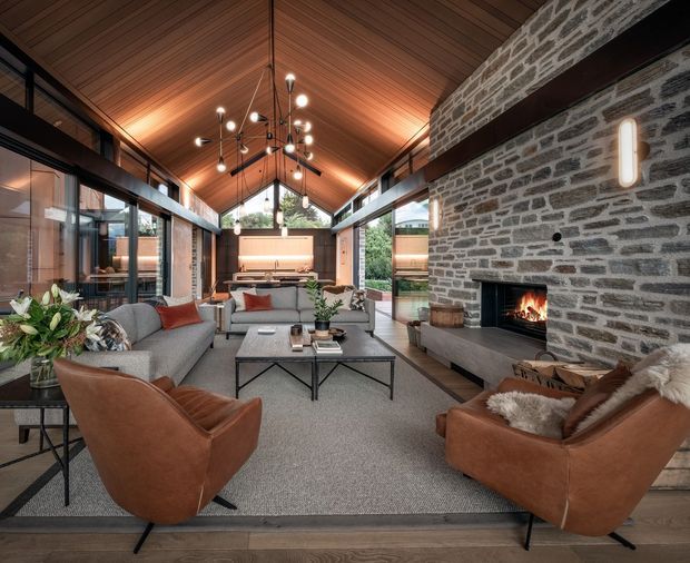 A sophisticated open-plan living room with a dramatic pitched timber-lined ceiling, exposed dark structural beams, full-height glazing, and a large exposed stone fireplace with a lit fire. The room features grey sectional seating, cognac leather lounge chairs, a dark metal coffee table, neutral carpeting, and sculptural pendant lighting. A kitchen or dining area is visible at the far end, while planted outdoor greenery is seen through the glazing.