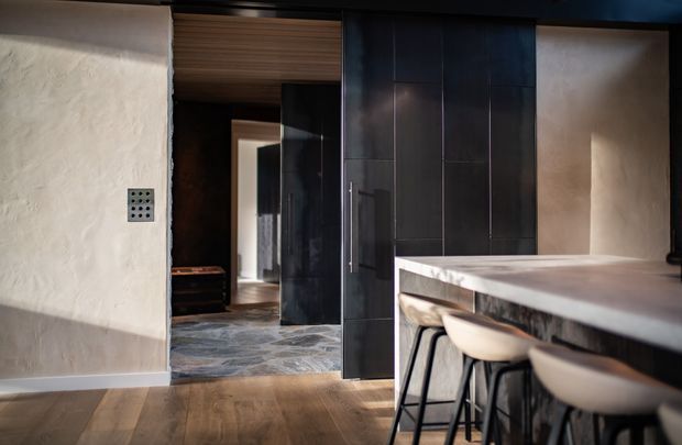 A moody contemporary kitchen interior viewed from an adjacent living or circulation area. The space features a dark, full-height tiled or panelled kitchen wall with integrated cabinetry, a pale stone island or peninsula with rounded light upholstered bar stools, textured off-white plaster walls, warm timber flooring, and a dark timber ceiling detail. A doorway and reflective dark surfaces extend the view into adjoining spaces.