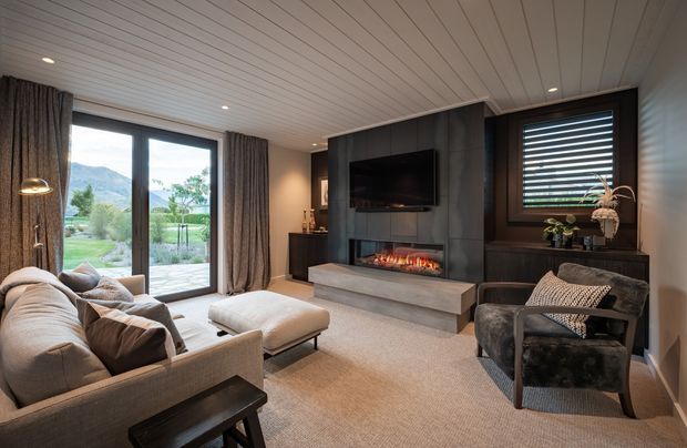 A refined contemporary living room with a large sectional sofa and ottoman, armchair, and dark built-in media wall centered on an electric fireplace and television. Full-height glazed doors open to a landscaped outdoor area and mountain view, while a slatted white ceiling, recessed lighting, dark timber joinery, layered curtains, and neutral furnishings create a warm, comfortable interior.