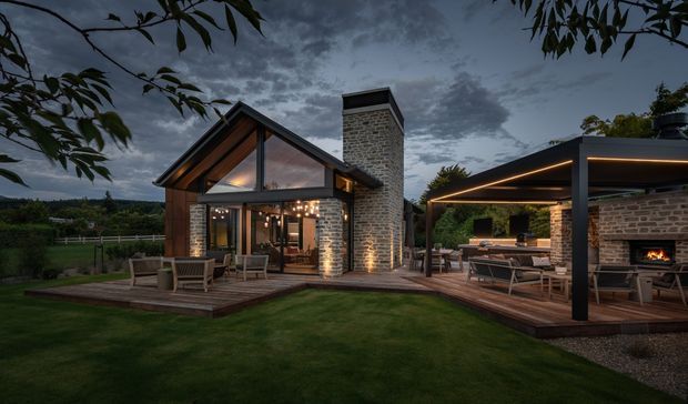 Twilight exterior view of a contemporary pavilion-style home set in a landscaped rural garden. The main structure has a steep gabled roof with exposed timber rafters, extensive glazing, dark-framed openings, and exposed light-grey masonry walls. A broad timber deck connects the house to a separate covered outdoor entertaining pavilion, which includes a built-in kitchen or bar area and an open fireplace. Warm integrated lighting highlights the roof edges, masonry, deck, and outdoor living spaces.