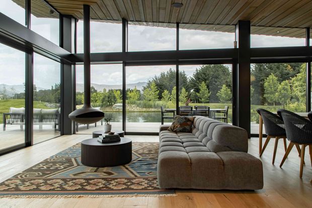 A spacious contemporary living room with floor-to-ceiling black-framed glazing overlooking a landscaped pond and trees. The room features a large curved tufted sectional sofa, patterned area rug, round ribbed coffee table, sculptural suspended fireplace, dining area, and a timber slatted ceiling. The interior has strong indoor-outdoor connection and abundant natural light.