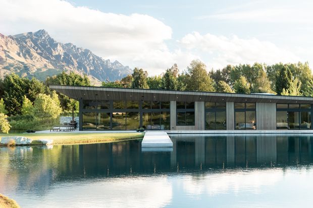 A low, elongated contemporary building with extensive floor-to-ceiling glazing and dark horizontal roof cladding sits beside a large reflecting pool. The architecture is set within a landscaped garden, with dramatic alpine mountains visible in the background. A small projecting platform extends into the water from the building.