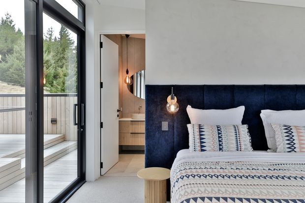 A contemporary bedroom with a deep navy upholstered floor-to-ceiling headboard, patterned bedding, white pillows, and a small light wood bedside table. Black-framed full-height sliding glass doors open onto a timber deck with steps and surrounding trees. An open doorway reveals a compact timber-joinery bathroom or vanity area with a pendant light.