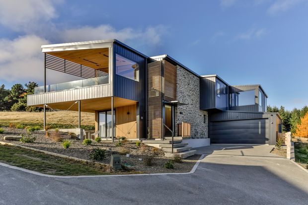 Contemporary multi-level residence on a sloping suburban site, combining dark metal cladding, extensive glazing, horizontal screening, natural timber soffits and a textured masonry feature wall. The home includes elevated balconies, a sheltered entry, landscaped gravel gardens and an integrated double garage.