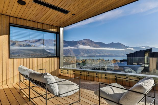 A modern covered outdoor lounge or sunroom with timber-lined walls, ceiling and decking, floor-to-ceiling glazing, and contemporary cushioned lounge chairs. The space overlooks a lake and dramatic mountainous landscape, with a glass balustrade and a dark-clad building visible outside.