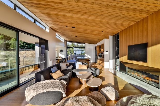 A bright, contemporary open-plan living room with a double-height or generously high sloping timber-lined ceiling, extensive floor-to-ceiling glazing, and warm timber flooring. The room features a dark sectional sofa, rounded coffee tables, soft textured throws and cushions, and a long built-in media wall with timber panelling, television, linear fireplace, and pale stone hearth. A kitchen and dining area are visible beyond the living space.