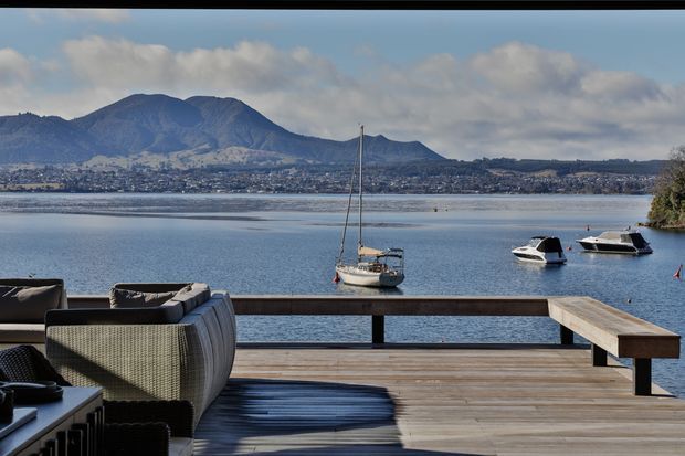 A waterfront outdoor deck overlooking a calm lake, with a timber railing and broad timber decking in the foreground. Dark woven outdoor lounge furniture occupies the left side, while several boats are moored on the water. A developed shoreline and prominent mountain range are visible across the lake beneath a partly cloudy sky.