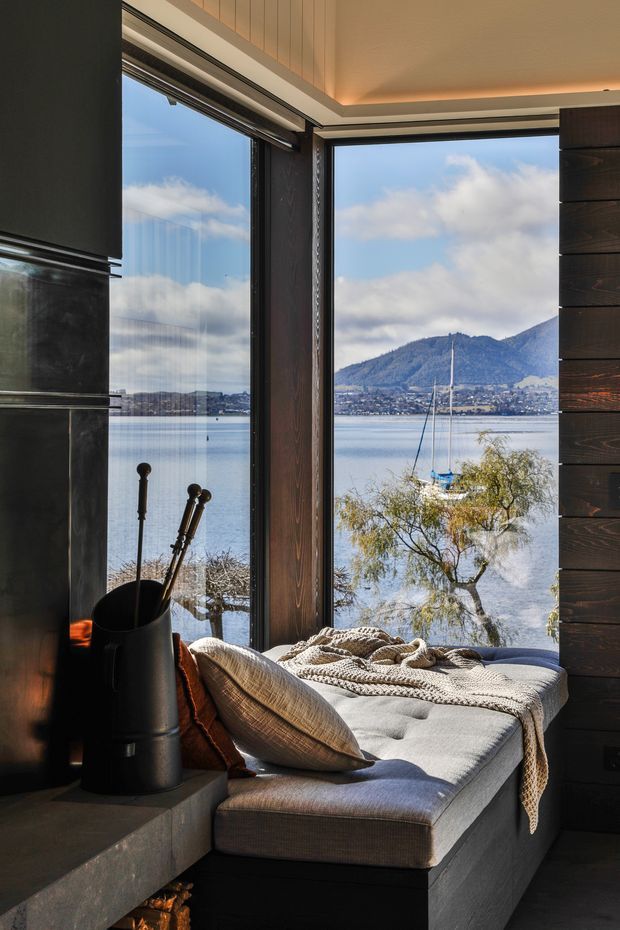 A cozy built-in window seat beside a dark fireplace, with a soft upholstered cushion, pillows and knitted throw. Floor-to-ceiling corner glazing overlooks a calm lake, sailboat and mountainous shoreline. The room features dark timber wall panelling, charcoal fireplace surfaces and warm concealed ceiling lighting.