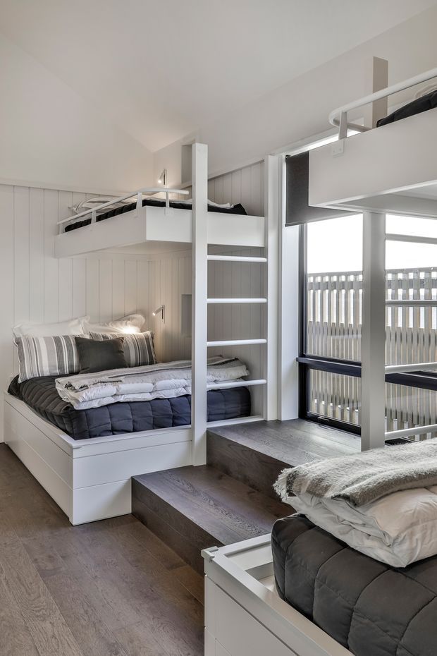 Bright, compact bunk-bedroom interior with two built-in white bunk beds and a lower bed, accessed by integrated ladder steps. White vertical wall panelling, dark timber flooring, and large windows with a balcony railing create a clean, contemporary coastal feel. Soft grey and white bedding adds a calm, practical atmosphere.