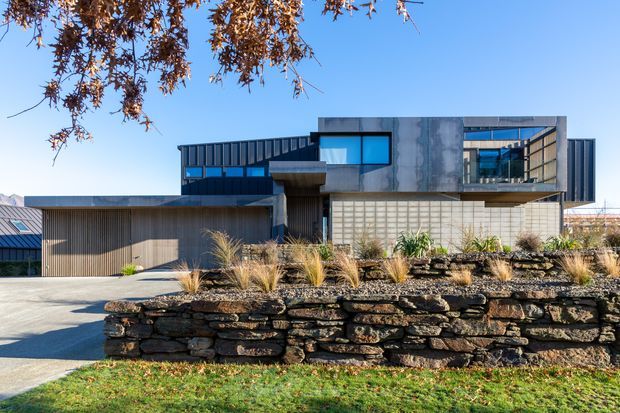 A contemporary multi-level residence viewed from the street, featuring a raw concrete upper volume, dark vertical metal cladding, extensive blue-tinted glazing, and a low horizontal garage or entry wing. A dry-stacked stone retaining wall and ornamental grasses form the landscaped foreground.