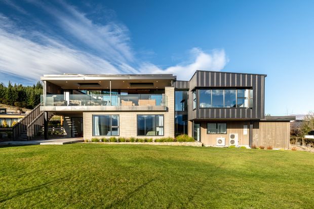 A contemporary multi-level residential house viewed from the rear across a broad lawn. The building combines pale masonry walls, dark vertical cladding, extensive glazing, a covered upper terrace with glass balustrades, and a flat roof. Exterior stairs connect the garden to the elevated living level, while the right wing features a tall glazed volume and timber-clad lower section.