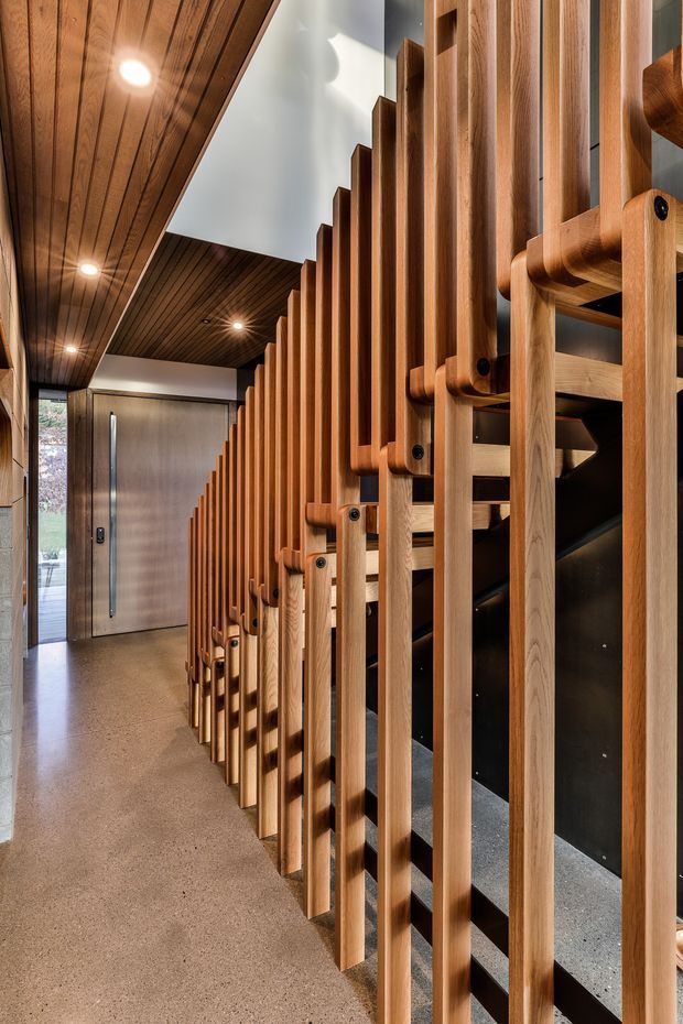 A refined residential entry hall featuring a dramatic sculptural timber screen or stair balustrade made from repeated vertical wood members, with integrated horizontal connectors and black fixings. The space has a dark timber-lined ceiling with recessed downlights, a polished concrete or terrazzo-like floor, dark wall surfaces, and a contemporary timber front door at the end of the corridor. Natural light enters from glazing near the entrance, creating a warm, architectural interior.