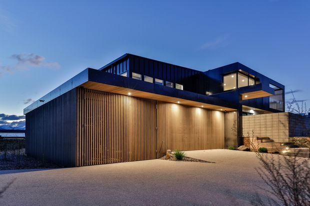 Exterior dusk view of a contemporary multi-level residence with a flat-roofed, angular composition. The facade combines dark vertical cladding, timber slatted screening, warm timber-lined soffits and walls, extensive glazing, and a pale masonry feature wall. Recessed exterior lighting illuminates the driveway and entry courtyard.