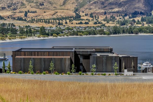 A low, contemporary lakeside building with dark vertical cladding, crisp geometric volumes, and flat rooflines. The structure sits beside a road and landscaped planting, with a marina or waterfront edge behind it and dry grass in the foreground. A broad lake and rugged, sparsely vegetated hills form the background.
