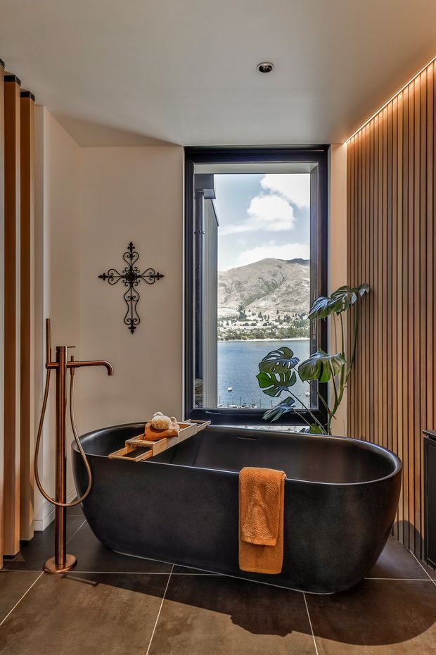 A refined bathroom featuring a freestanding matte-black bathtub, copper-toned floor-mounted tub filler and hand shower, dark tiled flooring, and a vertical timber slat feature wall. A large window frames a clear lakeside and mountainous view, while a potted monstera adds greenery. Warm concealed lighting highlights the timber wall.