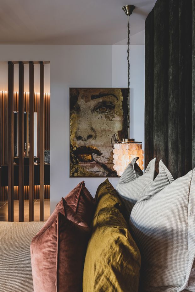 A moody, richly layered bedroom interior viewed close to the bed. Large textured cushions in blush, mustard, and light grey sit against the foreground bedding, with a sculptural pendant light, a large portrait artwork, dark floor-to-ceiling curtains, and a timber-and-dark-panel room divider with integrated lighting. The palette and lighting create a luxurious, dramatic atmosphere.