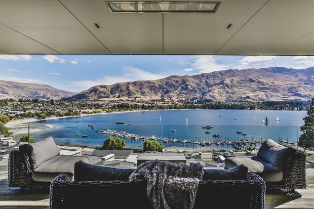 A covered outdoor lounge terrace overlooks a vivid blue lake, marina, and mountainous landscape. The space contains a large dark wicker sectional sofa with pale cushions, a furry throw, matching outdoor chairs, and low tables. A broad projecting roof or ceiling provides shade, creating a relaxed waterfront entertaining area.