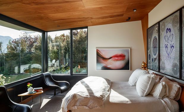 A bright, contemporary bedroom with a low-profile bed dressed in white textured bedding, two black leather lounge chairs, and a small timber side table. Floor-to-ceiling corner glazing opens to garden and mountain views, while the room features a warm timber-lined ceiling, dark window frames, neutral walls, and a large decorative patterned wall panel beside the bed. A framed artwork hangs above the bed area.