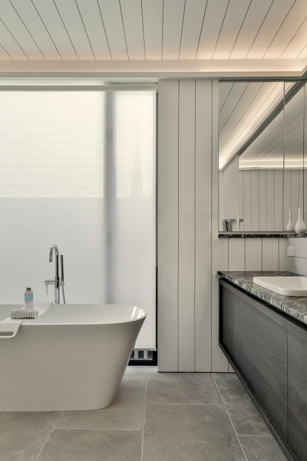 A refined contemporary bathroom featuring a freestanding white bathtub beside a large frosted window, a wall-mounted chrome bath filler, and a long dark timber-look vanity with a heavily veined stone benchtop and white vessel basin. Vertical light wall panelling, large grey floor tiles, a frameless mirror, and concealed linear lighting create a calm, minimalist composition.