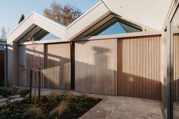 Contemporary low-rise building exterior with a dramatic folded, gabled roofline, pale horizontal cladding, and tall vertical timber screening panels. Triangular clerestory glazing sits beneath the roof peaks, while a stone-paved walkway borders a landscaped garden bed in the foreground.