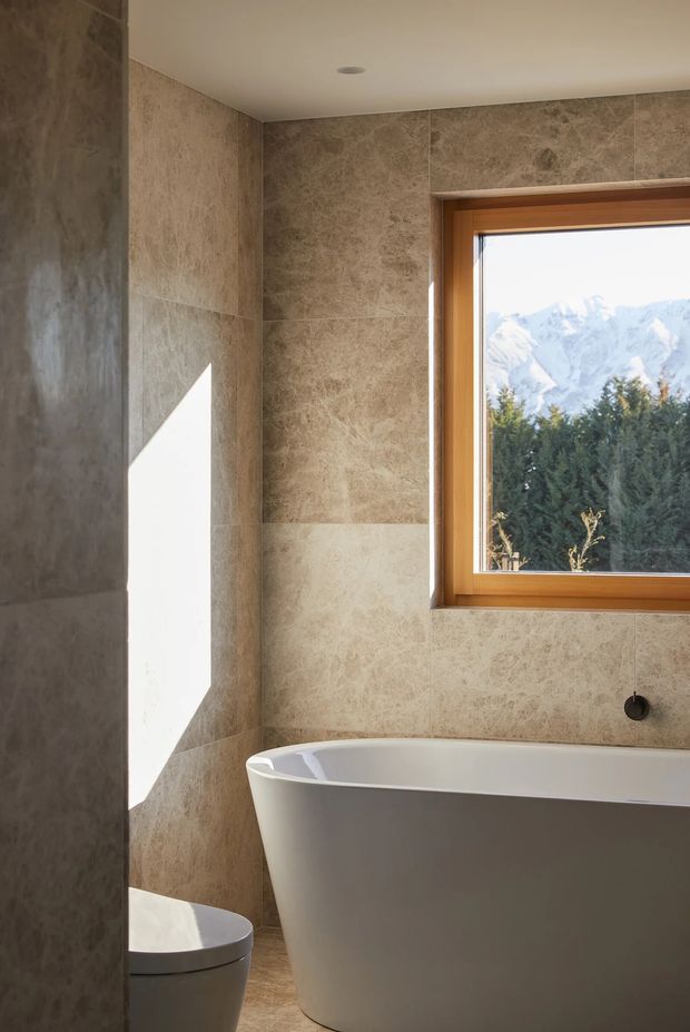 A warm, refined bathroom with full-height beige natural-stone-look wall tiling, a freestanding white bathtub, and a partially visible toilet. A timber-framed window provides bright natural light and overlooks dense greenery with snow-capped mountains beyond. The space has a simple smooth ceiling with a recessed downlight and a dark wall-mounted tap or fitting beside the bath.