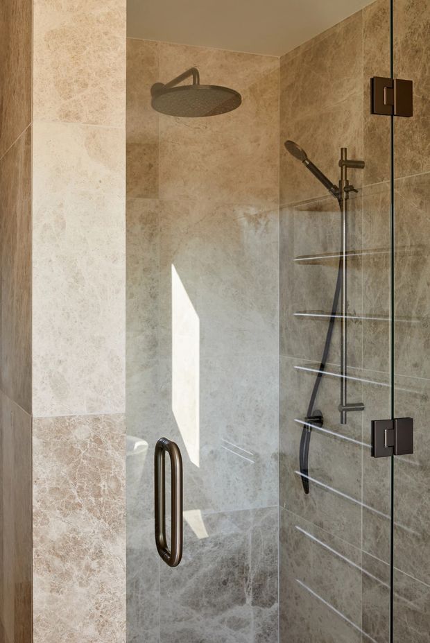 Close-up view of a contemporary walk-in shower enclosed by clear glass, with warm beige natural-stone-look wall tiles, a dark bronze rainfall showerhead and handheld shower, recessed glass shelves, and bronze-toned door hardware. Sunlight creates a bright geometric highlight across the shower wall.